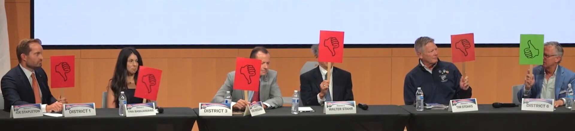 Six Newport Beach city council candidates sit at a table holding up vote cards. Joe Stapleton, Tina Babajanians, Erik Weigand, Walter Stahr and Tim Stoaks hold red thumbs-down cards. At the far right, behind the District 6 placard, Andrew Gerken holds a green thumbs-up card.