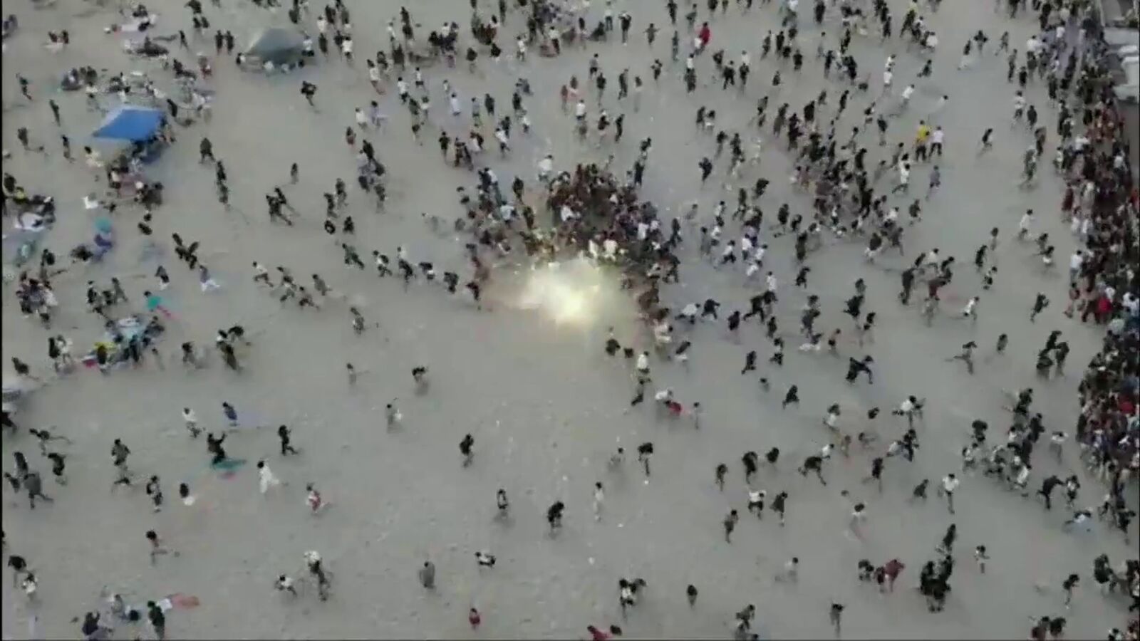 A frame from the city's drone video of the July 4, 2026 crowd on the sand near Newport Pier: hundreds of people running and scattering across the beach, with a bright flash at the center of the crowd.